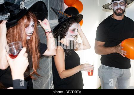 Amici adulti con cappelli da strega e trucco spettrale per il viso che si godono bevande e palloncini durante una divertente festa di Halloween al chiuso. Foto Stock