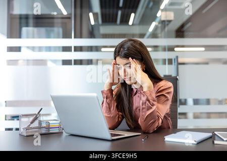 Donna d'affari che sperimenta un forte mal di testa e stress mentre lavora sul suo laptop in un ufficio moderno, sentendosi stanca e sopraffatta dalla pressione del lavoro Foto Stock