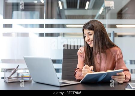 Giovane donna d'affari sorridente mentre scrive su un diario utilizzando un laptop, organizza compiti e pianifica a una moderna scrivania aziendale, concentrata, produttiva e sicura di sé Foto Stock