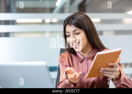 Giovane donna professionista che tiene una allegra videochiamata su un laptop, discute di lavoro e prende appunti in un quaderno durante una riunione remota in un ambiente di ufficio moderno Foto Stock
