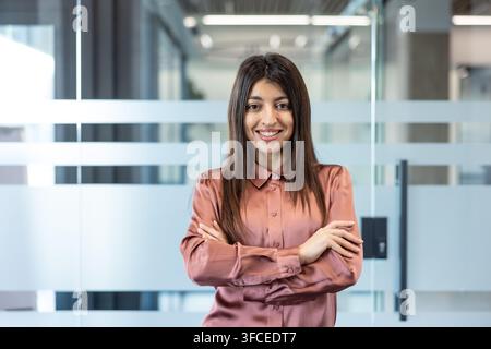 Giovane donna d'affari sicura di sé che posa in un ufficio moderno, rappresentando professionalità, successo e leadership nell'ambiente aziendale, offrendo un'immagine positiva e di empowerment Foto Stock
