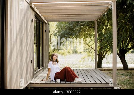 Una donna si siede comodamente su un terrazzo di legno, godendosi la vista serena sull'acqua e la vegetazione lussureggiante. Foto Stock