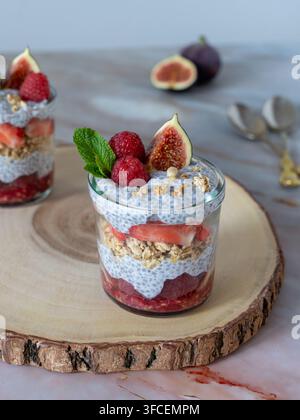 Dessert a strati individuale con panna bianca o yogurt, semi di chia, granola croccante, fichi freschi e lamponi, servito in un bicchiere su una fetta di legno rustico Foto Stock