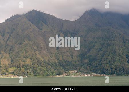 Le nuvole incombono sulle ripide pendici della caldera vulcanica e su un insediamento sul lago Batur a Kintamani, Bali. Foto Stock