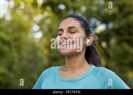 Giovane donna che sorride con gli occhi chiusi e indossa auricolari wireless, godendosi la musica mentre è in piedi in un parco con alberi verdi sfocati, esprimendo gioia, serenità e relax Foto Stock