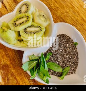 Vista dall'alto close-up di kiwi fresco affettato, semi di chia e foglie di menta verde serviti in un piatto di ceramica bianco a forma di cuore su sfondo di legno quadrato Foto Stock
