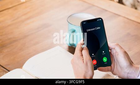 Primo piano di mani che tengono uno smartphone che mostra una chiamata in arrivo sconosciuta accanto a un quaderno aperto e una tazza di caffè su un tavolo di legno. Foto Stock