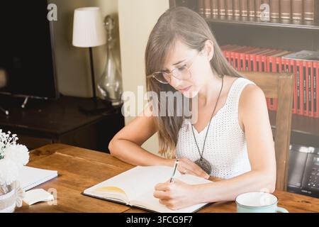 Giovane donna con occhiali scrive in un quaderno aperto su un tavolo di legno rustico, lavorando in silenzio in un accogliente ufficio domestico con scaffali di libri. Foto Stock