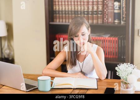 Giovane donna pensierosa con gli occhiali studia un quaderno aperto accanto a un laptop, uno smartphone e una tazza di caffè su una scrivania di legno in ufficio domestico. Foto Stock
