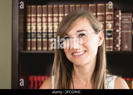 Ritratto ravvicinato di una giovane donna sorridente con occhiali davanti a una libreria, che esprime sicurezza e ottimismo a casa. Foto Stock
