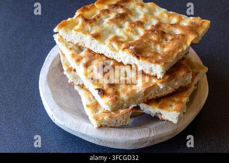 Fette di focaccia dorate impilate su una tavola rotonda di legno chiaro contro uno sfondo grigio scuro ruvido, angolo di 45 gradi Foto Stock