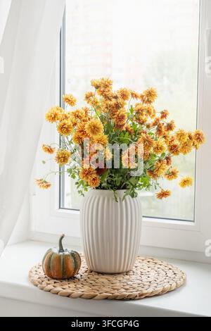 Mazzo di crisantemi arancioni in vaso bianco con piccola zucca su davanzale luminoso, decorazione autunnale stagionale per la casa con luce naturale. Formato verticale. Foto Stock