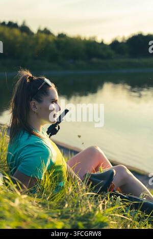 Giovane donna che riposa sul bordo dell'acqua del lago è seduta nell'erba con le ginocchia piegate e lo sguardo fisso davanti a sé. Una luce calda del tramonto sfiora il suo viso mentre riflessi calmi Foto Stock