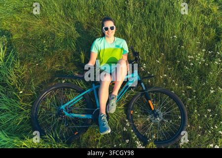 Ciclista sorridente seduta su una mountain bike in un prato verdeggiante, indossa occhiali da sole bianchi e una maglia sportiva. La sua posa rilassata Foto Stock