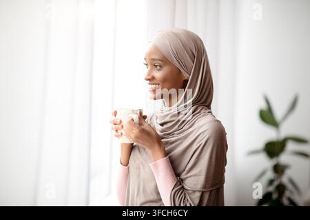 Attraente donna nera in hijab con una tazza di caffè caldo che guarda fuori dalla finestra di casa, gustando la sua bevanda calda. Bella signora millenaria nella headsca musulmana Foto Stock