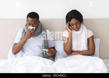 L'uomo e la donna di famiglia di colore hanno avuto il coronavirus, rimanendo a letto a casa. Coppia afroamericana ammalata seduta a letto, avvolta da sciarpe, con mal di testa e Foto Stock
