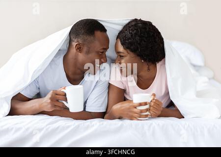 Amata coppia nera che si stende sotto la coperta, beve tè caldo, si riscalda durante il freddo. Amanti afroamericani felici con tazze che alloggiano a letto Foto Stock