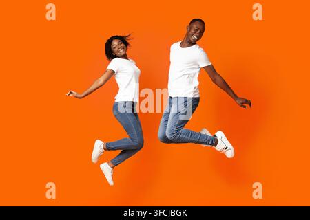 Giovane uomo afroamericano bello e donna carina che saltano sopra uno sfondo arancione in studio, con magliette bianche e jeans, gioiosamente neri Foto Stock