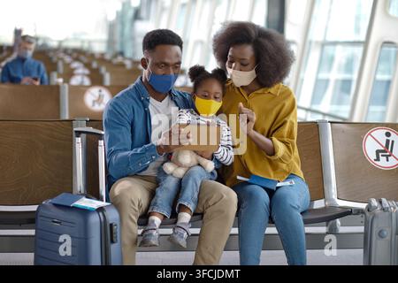 Un padre, una madre e una figlia sono seduti insieme in un terminal aeroportuale, tutti indossano coperture protettive per il viso e guardano un dispositivo digitale Foto Stock
