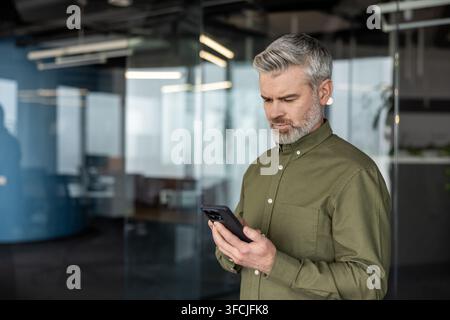 Uomo d'affari maturo in piedi, che guarda intensamente il suo smartphone e comunica utilizzando la tecnologia mobile in un ambiente aziendale contemporaneo con pareti di vetro Foto Stock