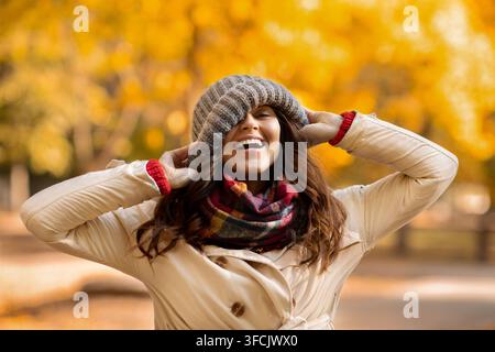 Giovane donna giocosa che si toglie il cappello in faccia e ride al parco nella calda giornata autunnale Foto Stock