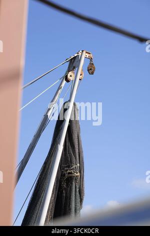 Rete da pesca sospesa da un braccio e una carrucola dell’imbarcazione, attrezzatura marittima dettagliata e sartiame logoro su una nave da pesca commerciale contro un cielo blu terso Foto Stock