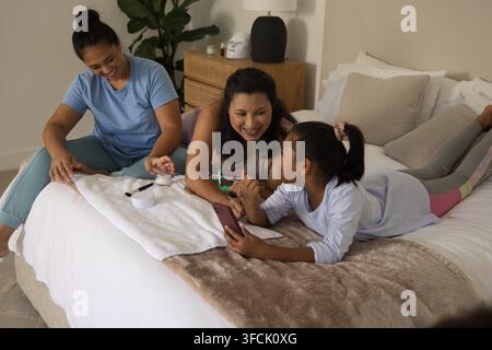 Diverse famiglia femminile seduta sul letto in una camera da letto illuminata dolcemente, con smartphone e vasetti di prodotti per la cura della pelle Foto Stock