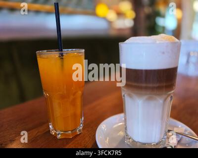 Un bicchiere di succo d'arancia ghiacciato con una cannuccia nera accanto a un caffè latte macchiato a strati servito su un tavolo di legno in un caffè Foto Stock