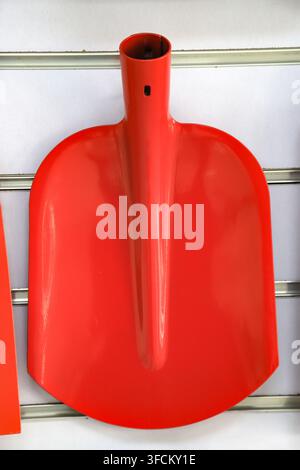 Immagine ad alta risoluzione in primo piano che cattura un singolo badile a testa quadrata arancione brillante ordinatamente appeso a un muro a doghe bianco in un negozio di ferramenta. Il troppo Foto Stock
