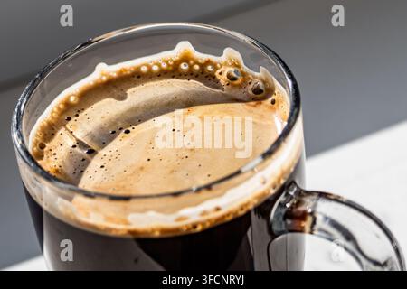 Macro di un espresso appena preparato con ricca schiuma dorata e minuscole bolle in una tazza di vetro, illuminato intensamente da una luce solare calda e diretta. Foto Stock