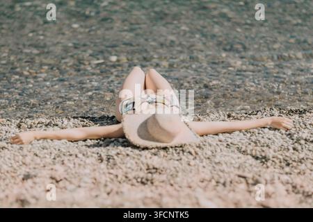 Donna che si rilassa in spiaggia, sdraiata su ciottoli ai margini dell'acqua, si fa abbronzare con un cappello di paglia e un bikini colorato, godendosi le vacanze estive Foto Stock