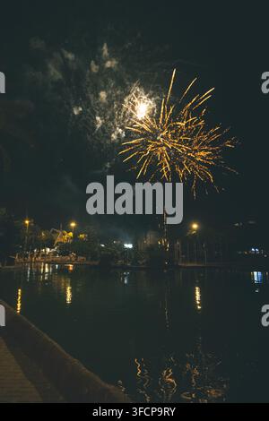 Fuochi d'artificio dorati vibranti illuminano il cielo notturno sopra un lago urbano tranquillo, creando un'atmosfera festosa e celebrativa in un ambiente di parco pacifico. Foto Stock
