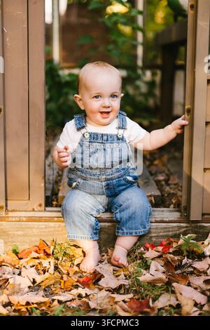 Bambino felice in tuta seduto tra le foglie autunnali Foto Stock