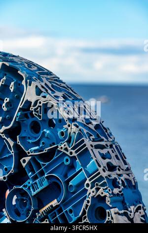 una grande scultura circolare realizzata con parti meccaniche blu e grigie stratificate, ambientata su uno sfondo sfocato di mare e cielo Foto Stock