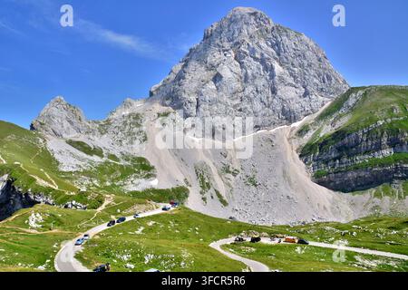 Un drammatico picco di calcare si erge su una strada tortuosa del passo montano con auto parcheggiate, circondato da prati alpini verdi sotto un cielo blu terso. Foto Stock