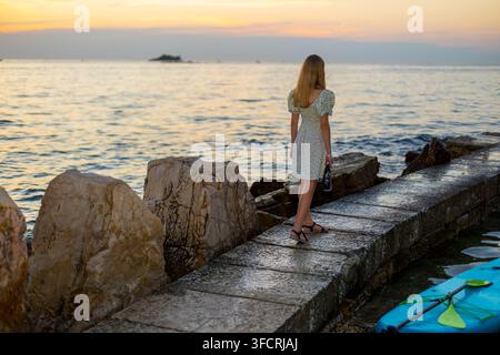 Ragazza alla moda in piedi su un molo al tramonto. Ragazza alla moda in un vestito floreale in piedi su un molo di pietra vicino al mare al tramonto, con rocce costiere e Foto Stock