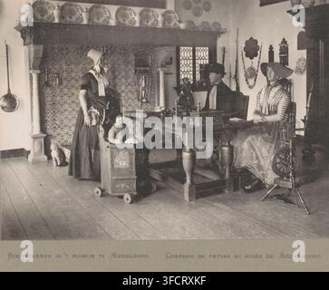 Camera di un contadino nel museo di Middelburg, Chambre de Paysan au musée de Middelbourg, Camera di un contadino in un museo a Middelburg, stampa fotomeccanica, produttore: Straaten, Gebroeders van, Middelburg, c. 1890 - c. 1910, carta, collotipia, fotogravura, altezza 156 mm x larghezza 216 mm, contadini, esposizione permanente, museo, costume folkloristico, costume regionale., Una scena d'interni in toni di seppia mostra una stanza con assi di legno sul pavimento e piastrelle murali a motivi sotto una grande mensola del camino che espone piatti e vasi decorativi; sotto il camino una donna in abito tradizionale e cappello si trova accanto a un bambino piccolo Foto Stock
