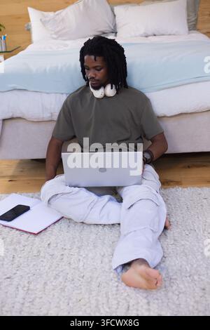Uomo sulla ventina seduto su un tappeto in camera da letto che usa un laptop con cuffie, smartphone, quaderno nelle vicinanze Foto Stock