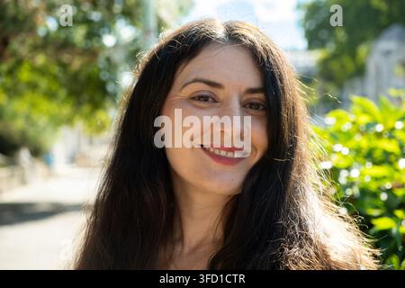 Donna adulta felice con lunghi capelli scuri che sorride alla luce naturale all'aperto. Foto Stock