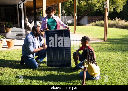 Famiglia afroamericana che assembla un pannello solare sul prato del cortile illuminato dal sole vicino a una pergola Foto Stock