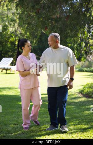 Caring asiatica in tuta rosa, anziano afroamericano che cammina sul prato tenendo l'avambraccio Foto Stock