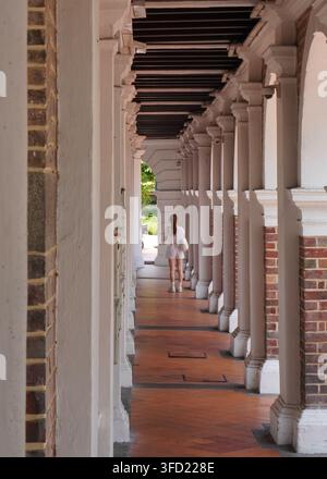 Donna che cammina da sola attraverso un corridoio del patrimonio coloniale. Vista verticale di una donna in bianco che cammina attraverso un lungo corridoio ad arcade in stile coloniale con r Foto Stock