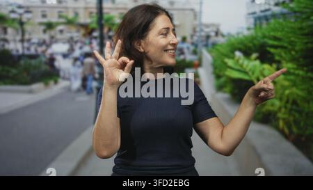 Una donna di mezza età fa un segnale ok con la mano sinistra e punta il dito destro su strade urbane trafficate; gesto di approvazione della fiducia. Foto Stock