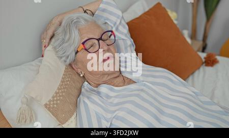 Donna anziana con occhiali colorati che dorme sul letto con la mano dietro la testa e supporto di cuscino in edificio; recupero nella serenità. Foto Stock