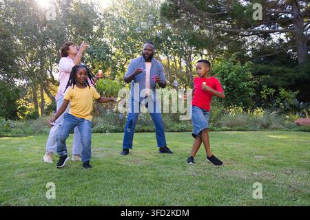 Famiglia afroamericana in abiti colorati che soffia e insegue bolle di sapone nel cortile con le bacchette Foto Stock
