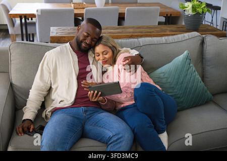 Coppia diversa che si appoggia insieme e guarda un tablet sul divano in salotto con smartphone Foto Stock