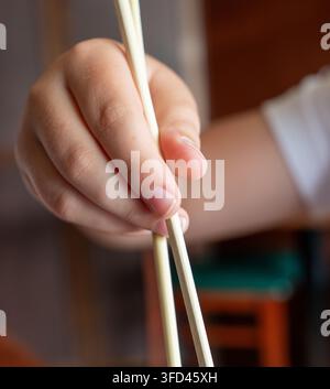 Primo piano verticale di una mano che tiene delle bacchette di legno mentre si cena in un ristorante di sushi. Una profondità di campo ridotta mette in evidenza le dita e le bacchette Foto Stock