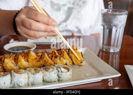 Prima piano di una persona che usa delle bacchette di legno per prendere un rotolo di sushi fritto e croccante da un piatto bianco in un ristorante giapponese. Salsa di soia e un bicchiere o Foto Stock