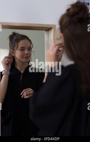 Donna che regola un orecchino a cerchio, controlla lo specchio in una camera da letto con cornice in legno, indossa una vestaglia nera e un orologio Foto Stock