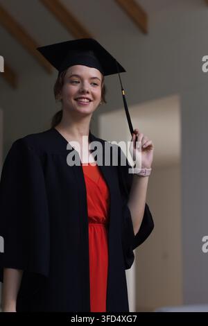 Laureata che tiene il nappino della toga mentre è in piedi in una stanza, indossa una toga nera e un vestito rosso Foto Stock